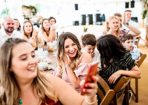 Wedding guests smiling and taking selfies at a festive outdoor marquee reception.