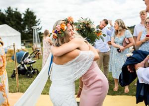 Bride in white lace dress with floral crown sharing a warm hug with a guest in pink at an outdoor summer wedding.