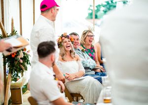 Bride wearing floral crown laughing joyfully during outdoor wedding ceremony under tent.