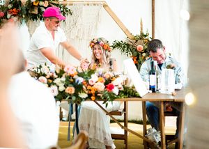 Bride in flower crown and groom signing wedding register at decorated boho ceremony table.