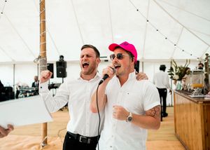 Two men in white shirts energetically singing into microphone at a wedding reception.