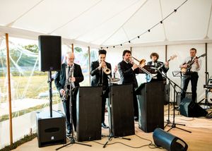 Live wedding band with brass section performing under a white marquee tent at a reception.