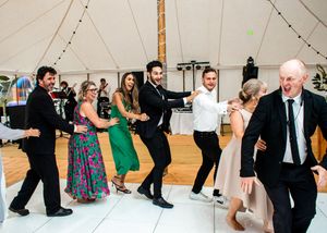 Wedding guests dancing joyfully in a line on the dance floor inside a white marquee tent.