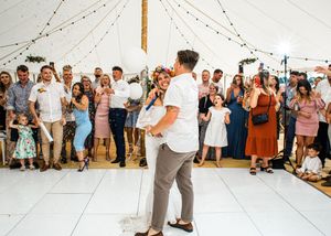 Bride and groom share romantic first dance surrounded by smiling wedding guests in marquee.