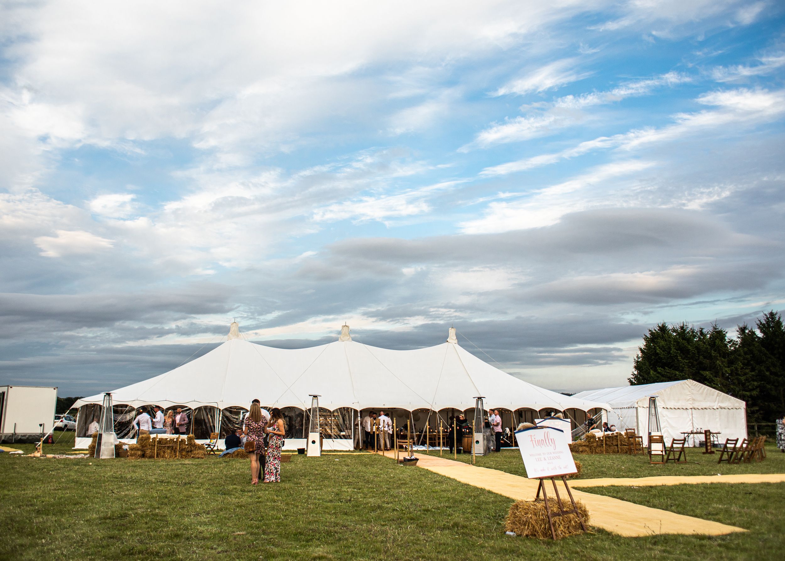 Large white wedding tent on green grass under dramatic cloudy sky with guests gathered outside.
