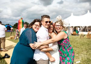 Four wedding guests laughing and hugging together on a sunny outdoor wedding venue with a white marquee tent.