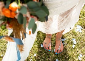 Close-up of brides feet in blue sandals with turquoise nails standing on grass holding an orange floral bouquet.