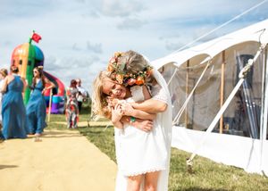 Bride in white lace dress and floral crown sharing a joyful embrace with a young child at an outdoor wedding.