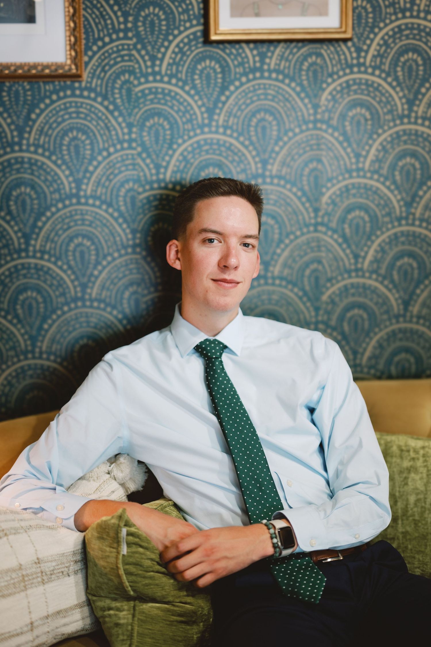 A person in a light blue dress shirt and green polka dot tie reclines on an olive green couch against patterned wallpaper.