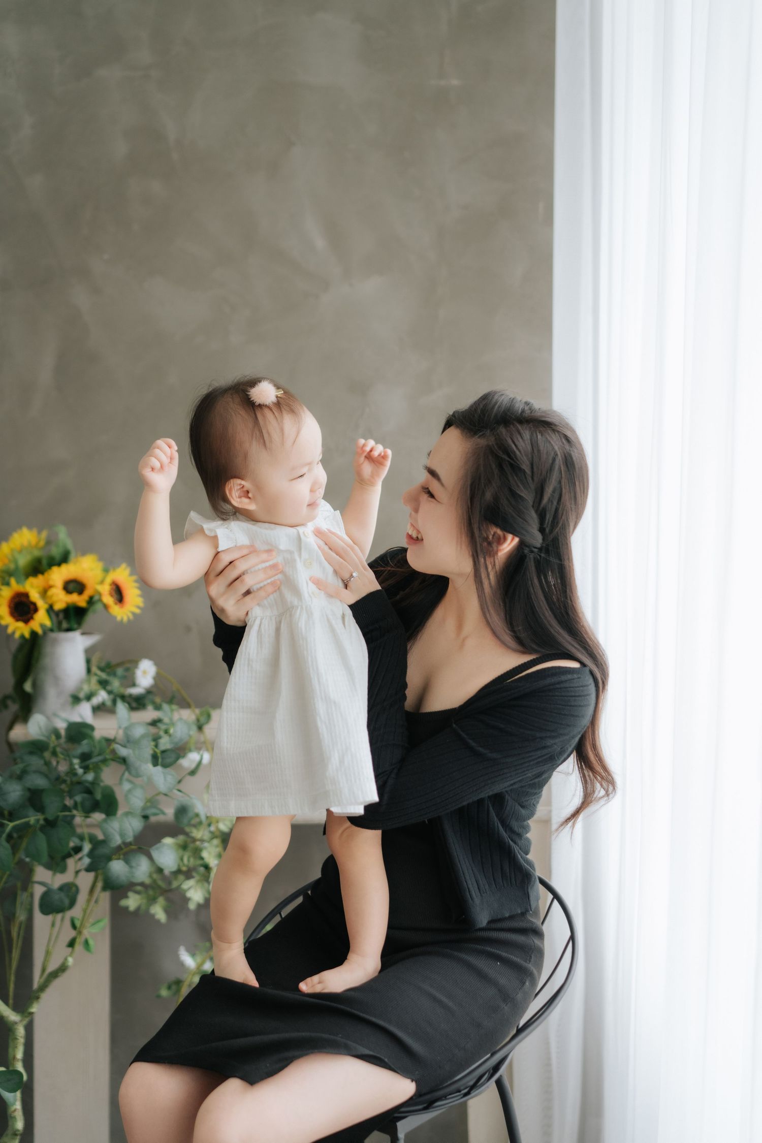 Mother and baby in white dress share a joyful moment by window with sunflowers and greenery in background.穿著白色洋裝的母親和嬰兒在窗邊享受快樂的時光，背景是向日葵和綠植。
