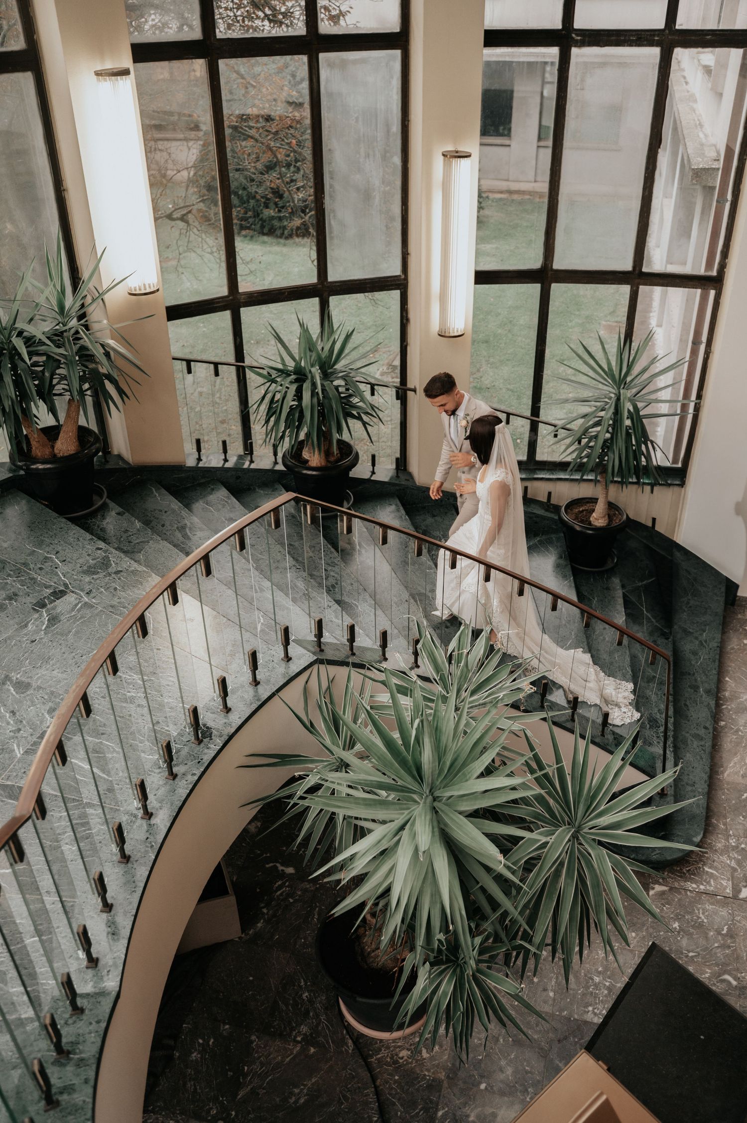 A bride and groom stand on an elegant curved marble staircase surrounded by tropical plants in a grand windowed venue.