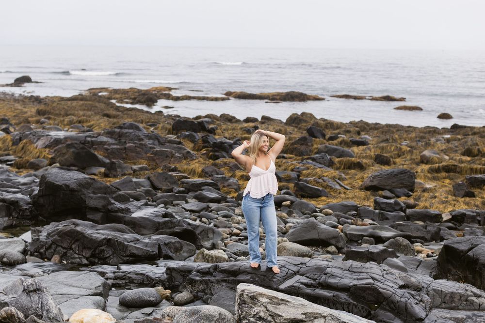 Landscape photo of blond girl wearing a white tank and blue jeans poses on dark coastal rocks for senior photos at North Hampton Beach NH