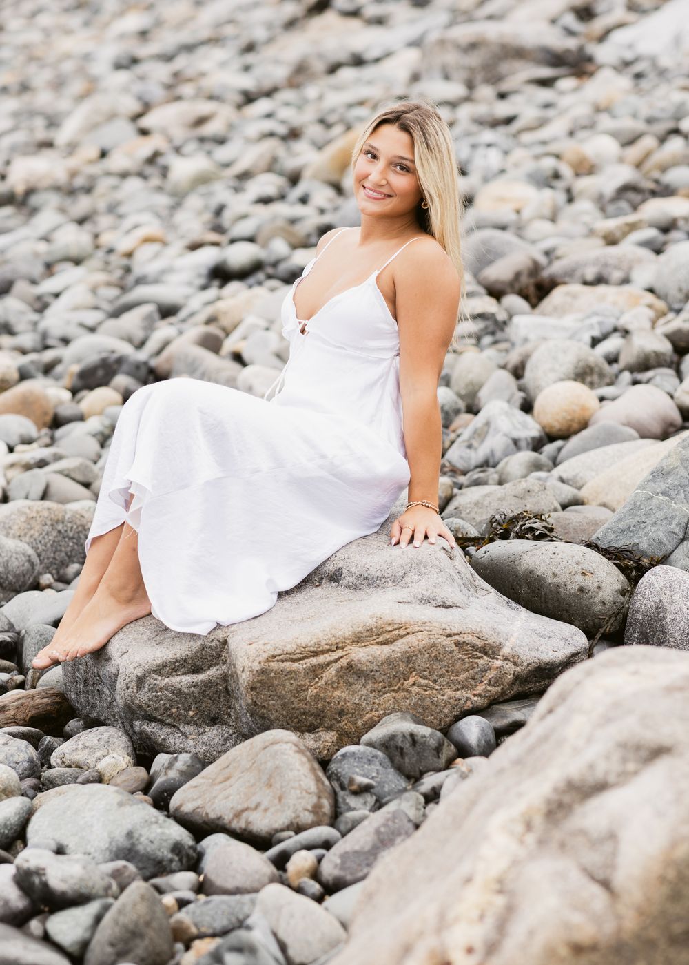 Blond girl sits on light colored rocks on a beach wearing a white maxi dress for senior photos at North Hampton Beach. Seacoast senior photo