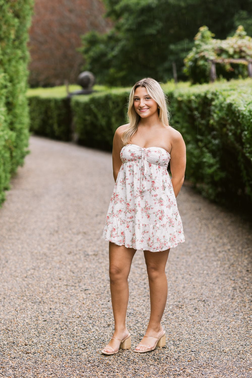 Blond senior girl wears a white floral dress stands on a gravel path at Fuller Gardens North Hampton, New Hampshire for senior photos.