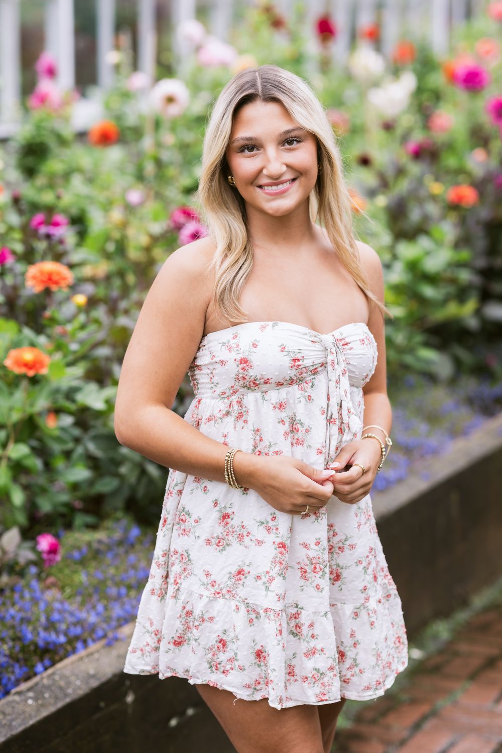 A blond girl wearing a short, white floral dress poses standing in front of Dalias for senior photos at Fuller Gardens in North Hampton, NH