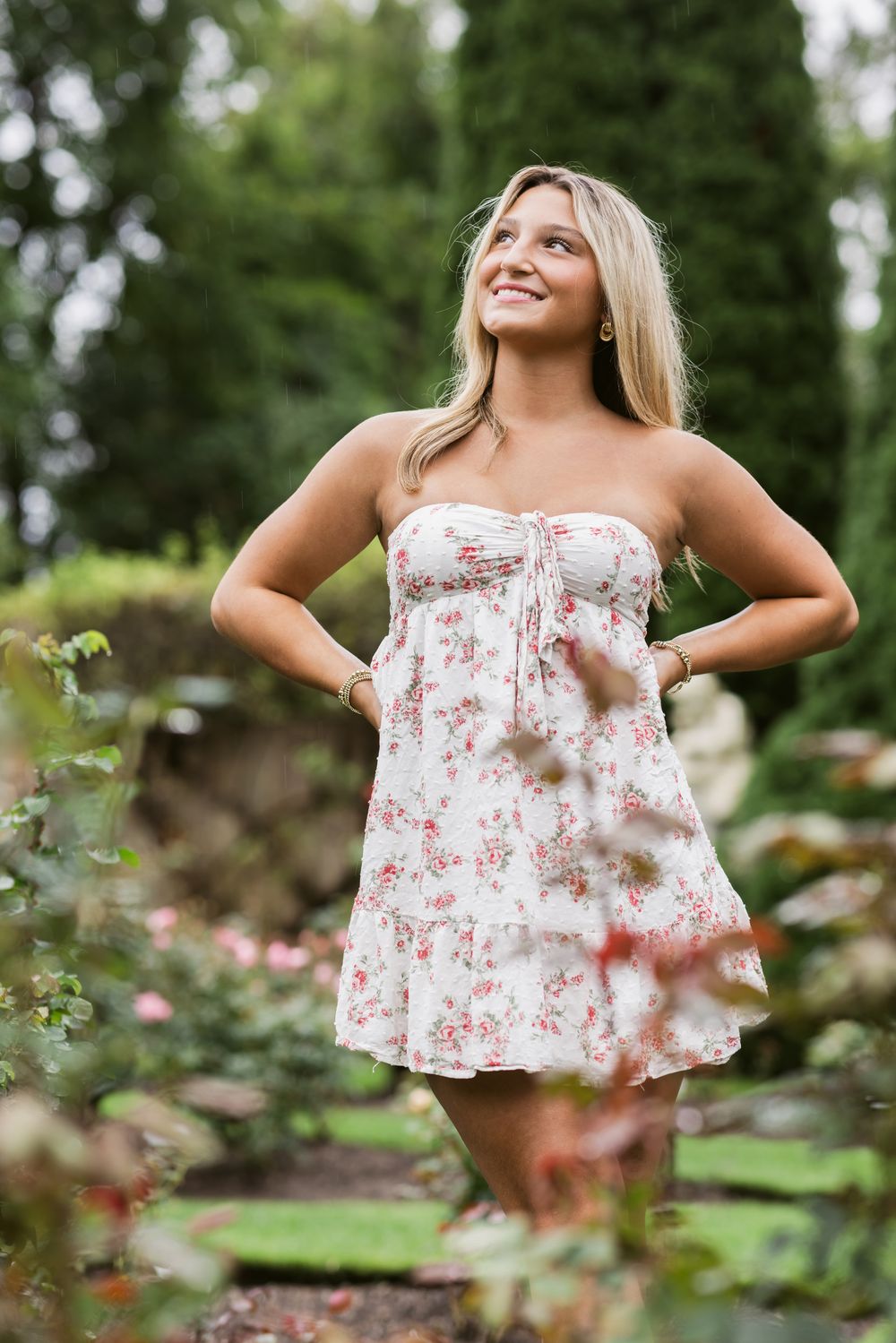 Senior girl poses with hands on her hips wearing a white short dress in a rose garden at Fuller Gardens in North Hampton, New Hampshire.