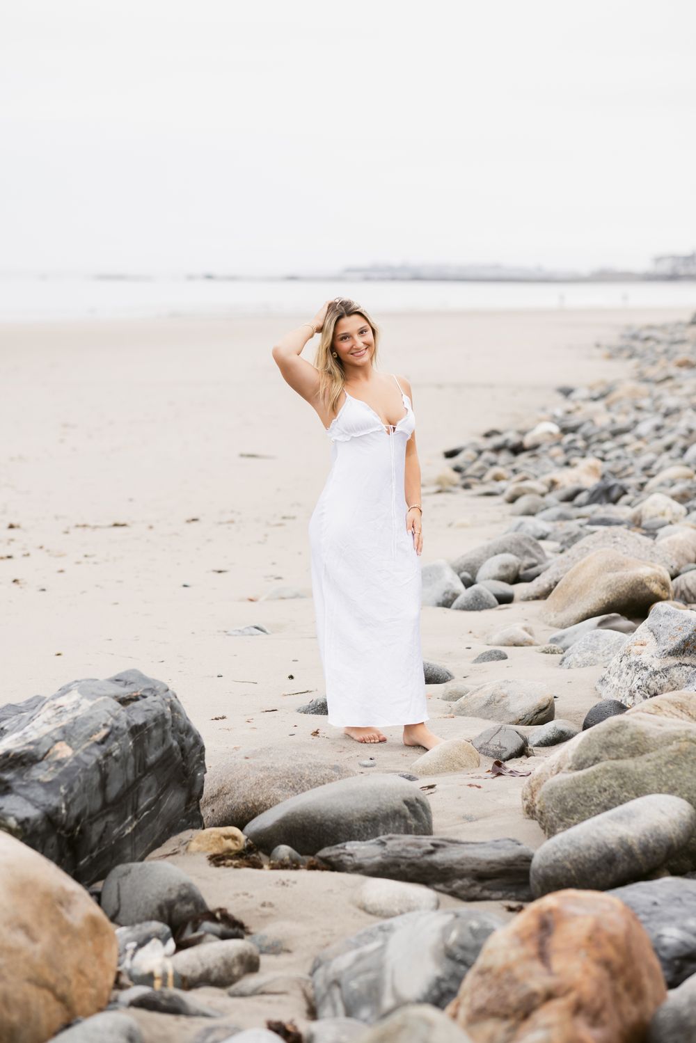 Girl poses on beach on a cloudy day wearing a long white maxi dress for senior photos at North Hampton Beach. Cohasset senior photographer.