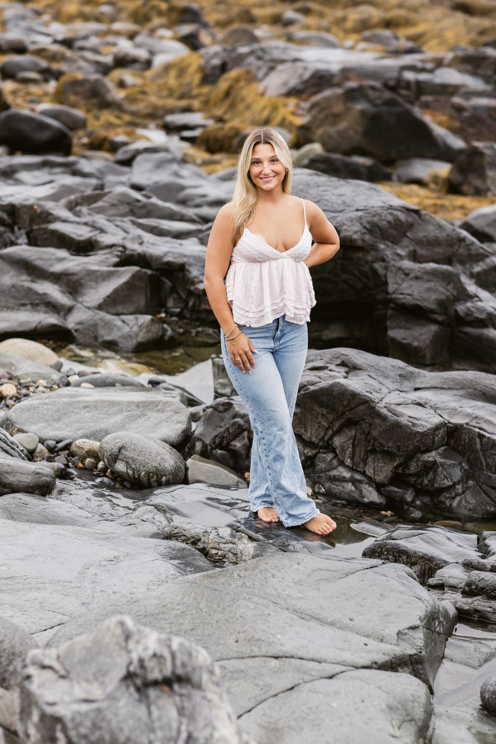 Blond girl stands on dark coastal rocks at North Hampton Beach for senior photos wearing white tank.Southern Maine York senior photographer