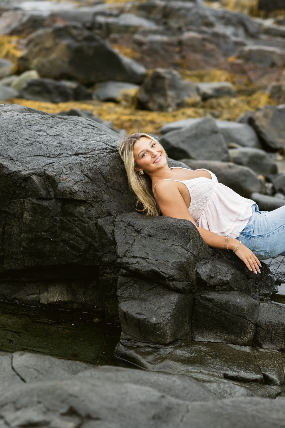 Blond senior girl wearing a white tank top and light blue jeans lays on dark coastal rocks for senior photos at North Hampton Beach NH.