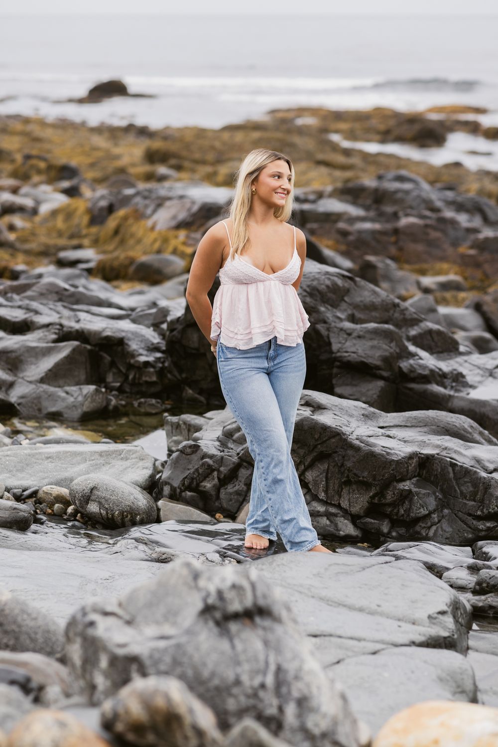 Blond girl stands on dark coastal rocks on cloudy day for senior photos at North Hampton Beach. North Shore Essex senior photographer.