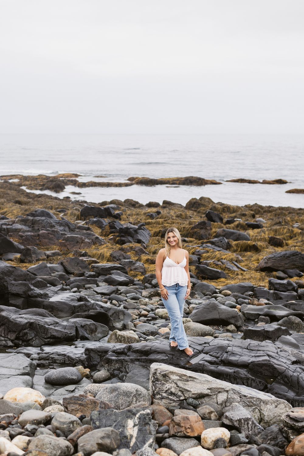 Landscape of blond girl wearing white tank and blue jeans poses on dark coastal rocks at North Hampton Beach. Gloucester senior photographer