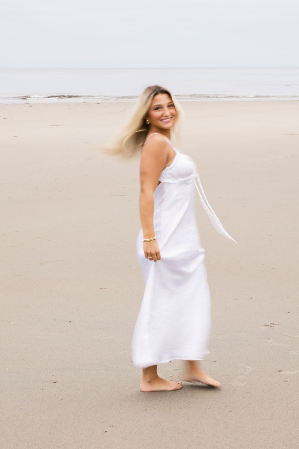 A slow motion photo of blond girl spinning wearing a long white maxi dress for senior photos at North Hampton Beach. Newburyport senior pics
