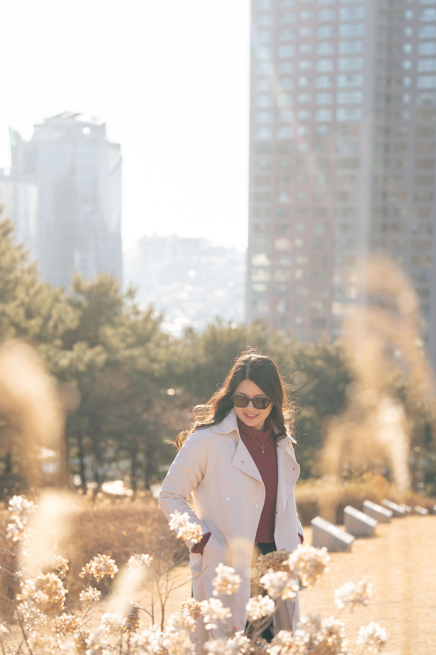 A person in a white blazer and sunglasses stands in a field of tall grass at golden hour with city buildings behind them.