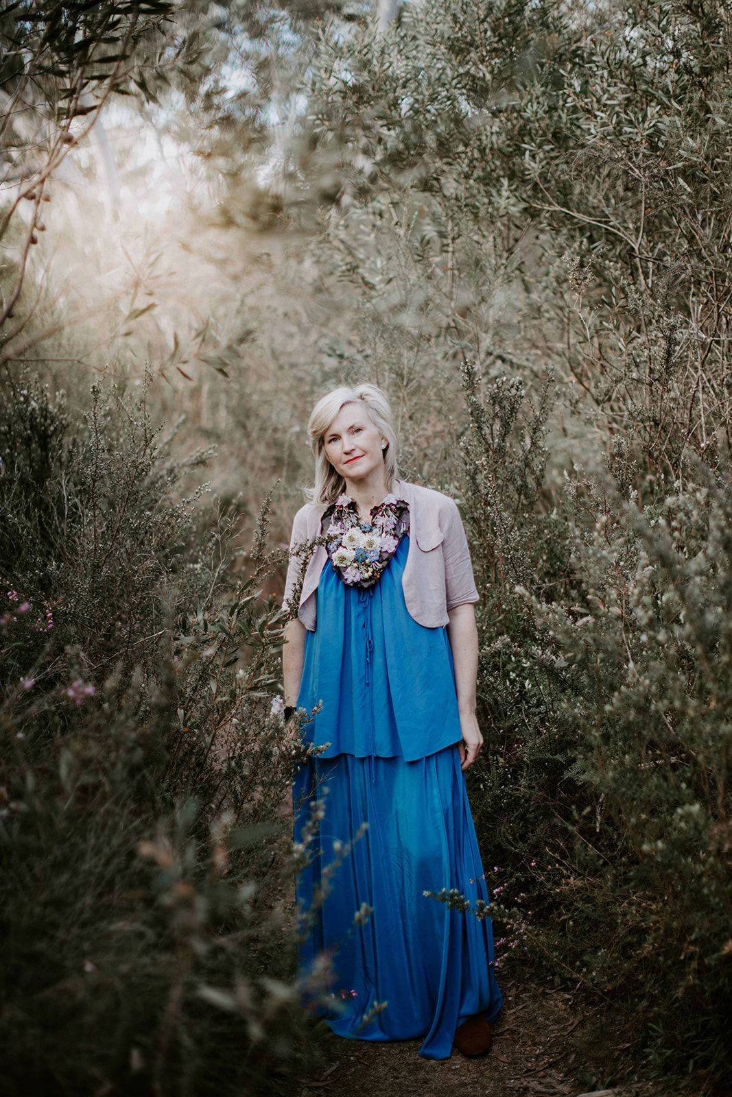 A person in a flowing blue dress and gray cardigan stands in a dreamy outdoor setting with natural foliage.