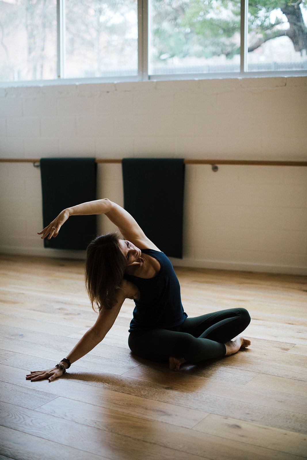 A dancer performs a graceful floor sequence in black attire on a wooden studio floor with natural light streaming in.