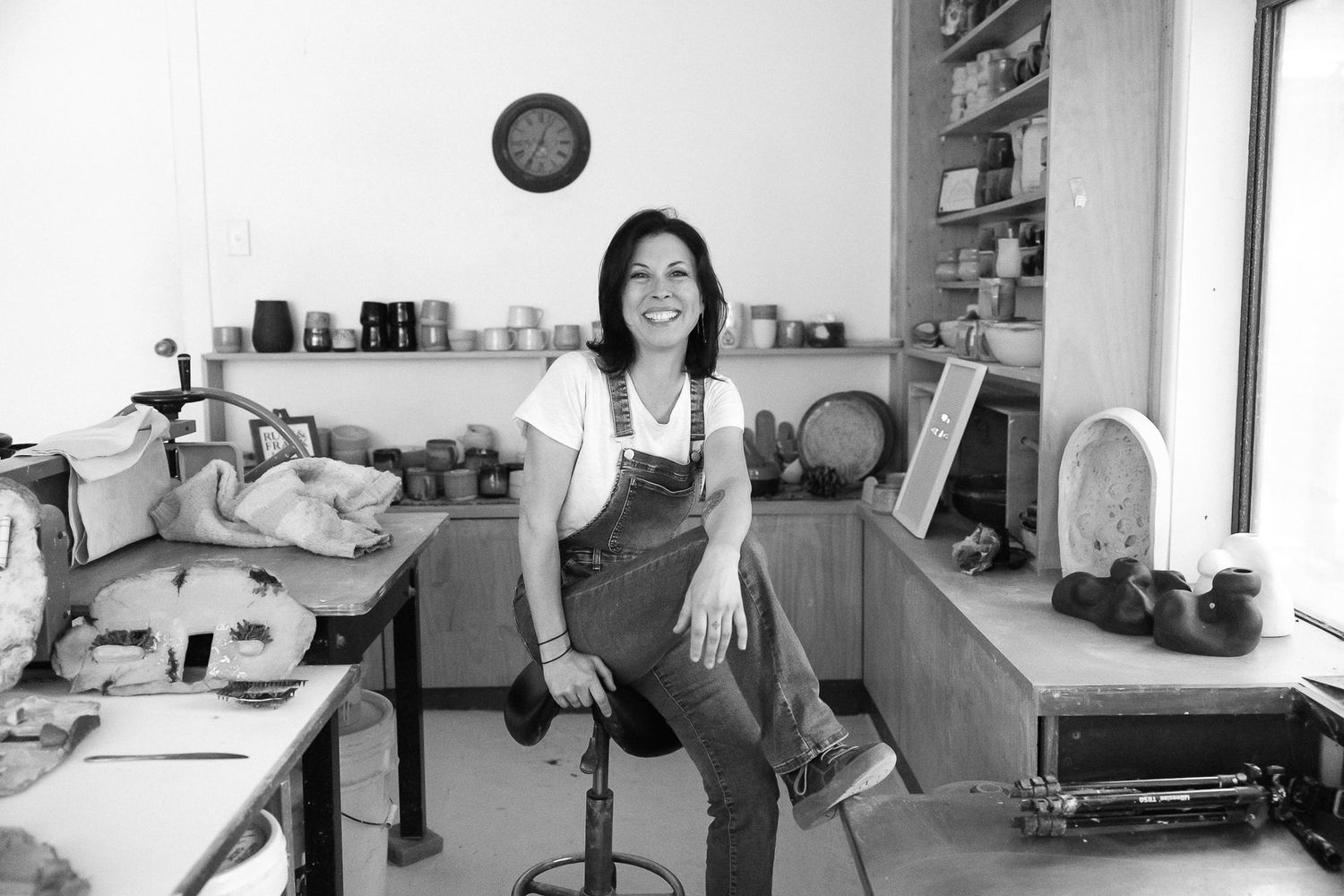 Woman smiling in a pottery studio workshop, surrounded by ceramic pieces and shelves, black and white photo.