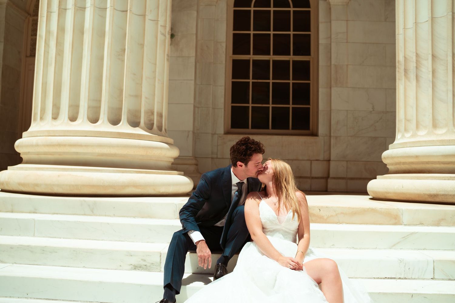 Bride and groom share a romantic kiss on marble steps surrounded by elegant white columns of a grand building.