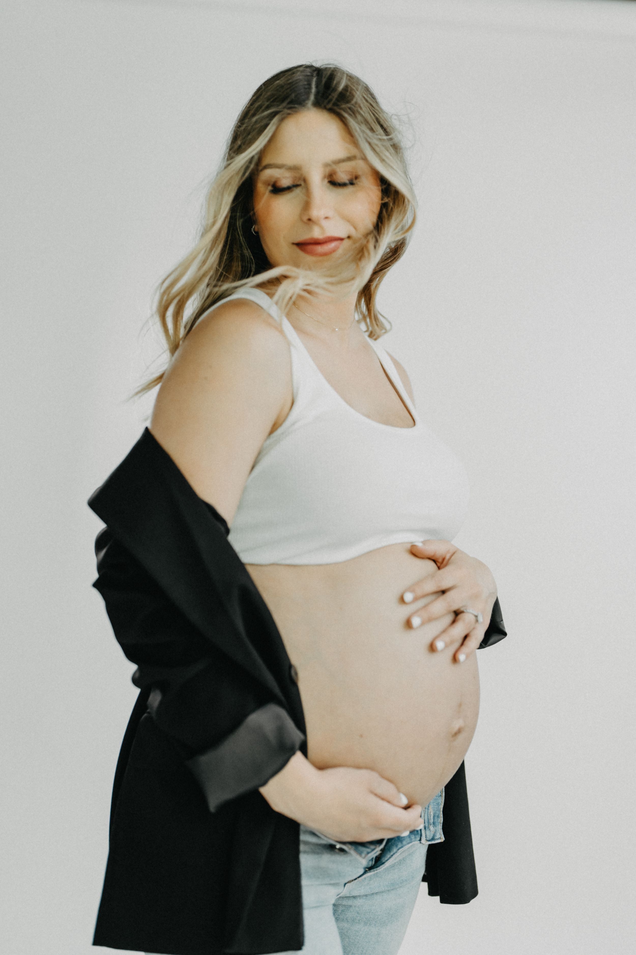 A smiling pregnant woman in a white tank top and black jacket cradles her baby bump in a studio.