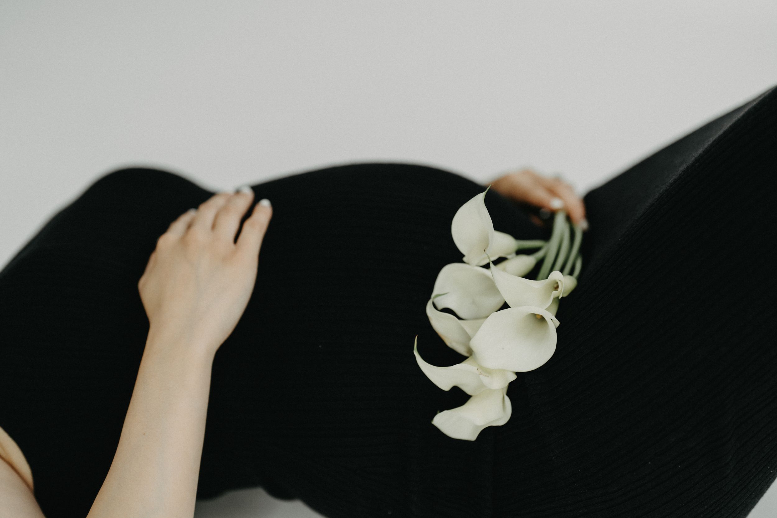 Close-up of pregnant belly in black dress holding white calla lilies.