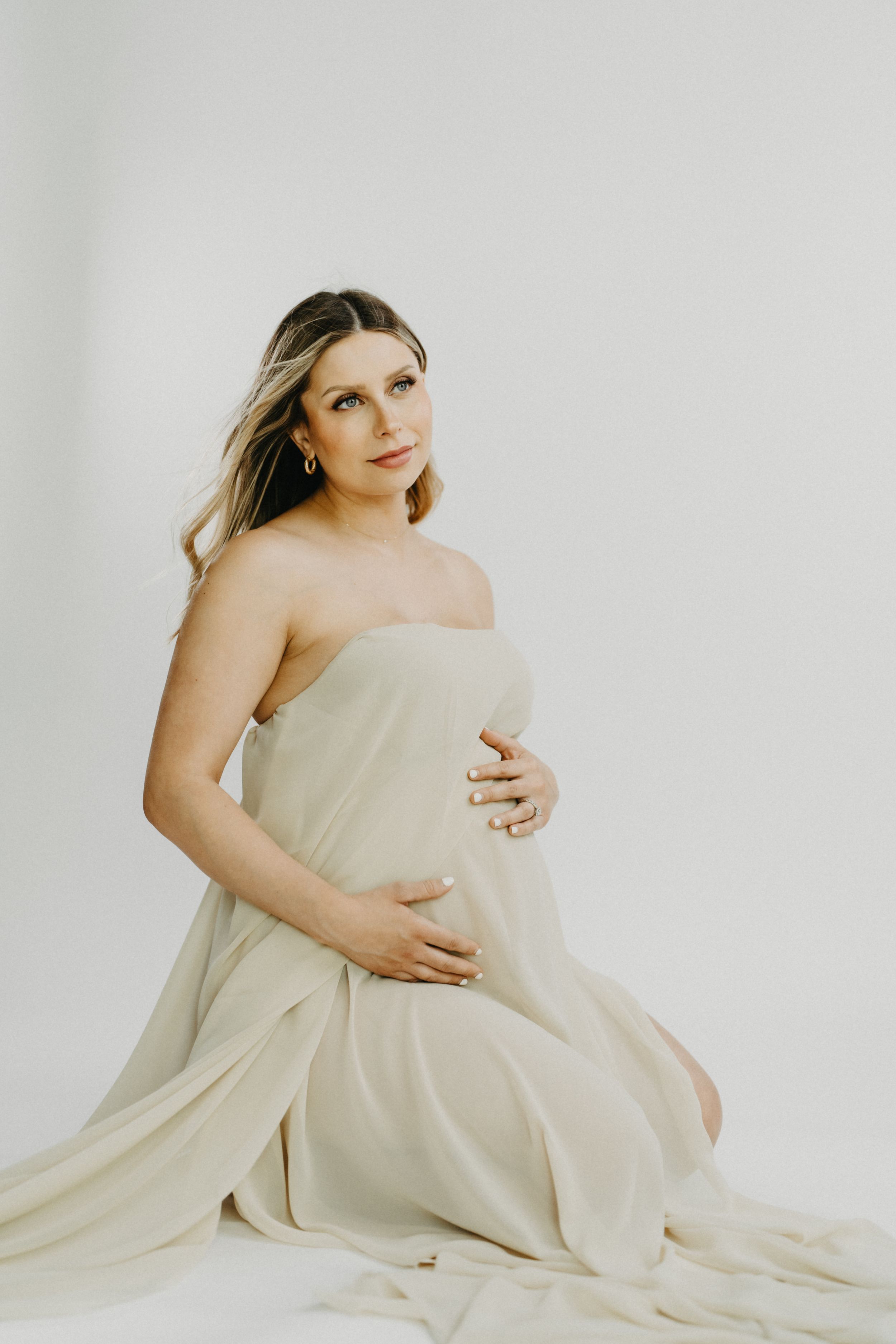 Pregnant woman wrapped in cream fabric sitting gracefully on white backdrop.