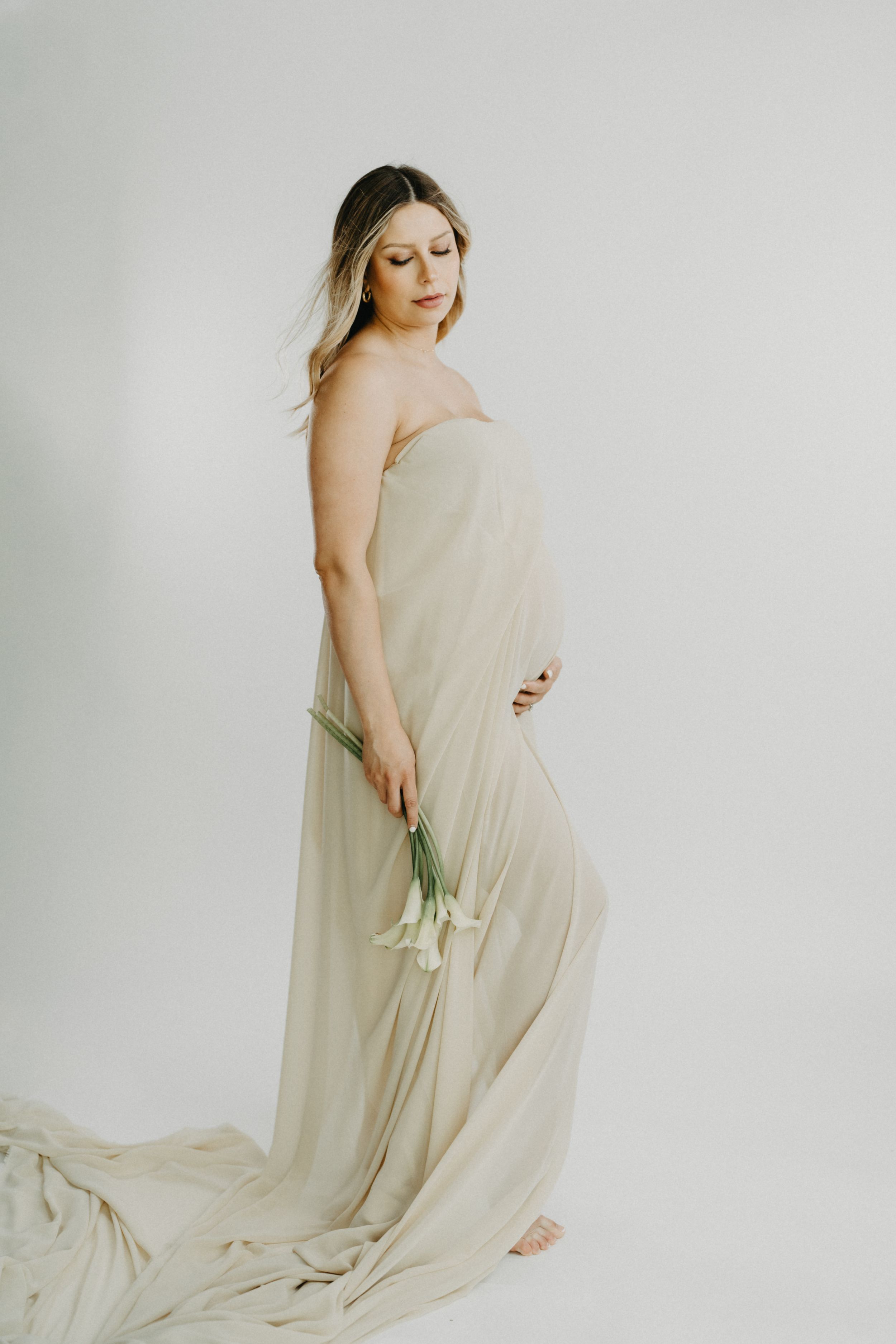 A pregnant woman draped in flowing beige fabric holds white flowers in a bright minimalist studio.