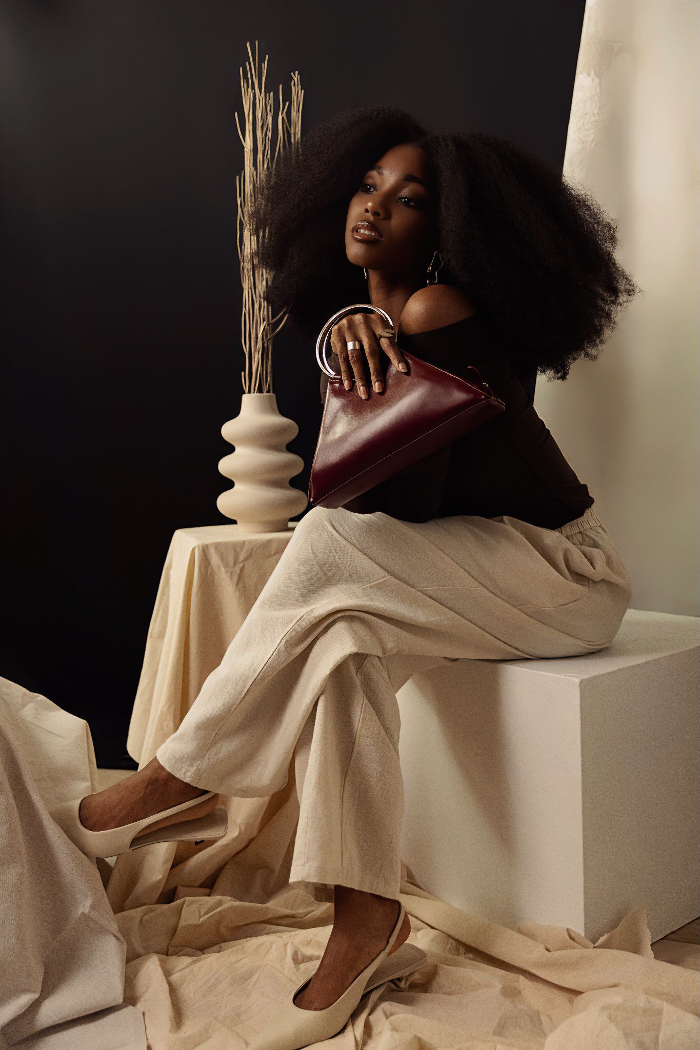 Woman with afro hair holding burgundy handbag seated on white cube in editorial studio setting.