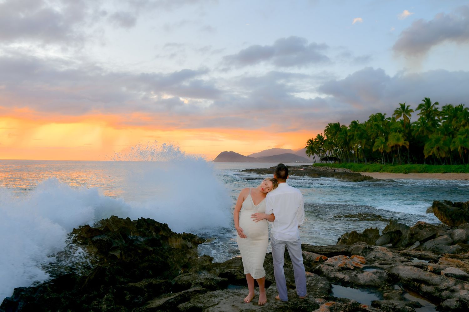 A couple watches ocean waves crash against rocky shore during vibrant sunset at tropical beach with palm trees.