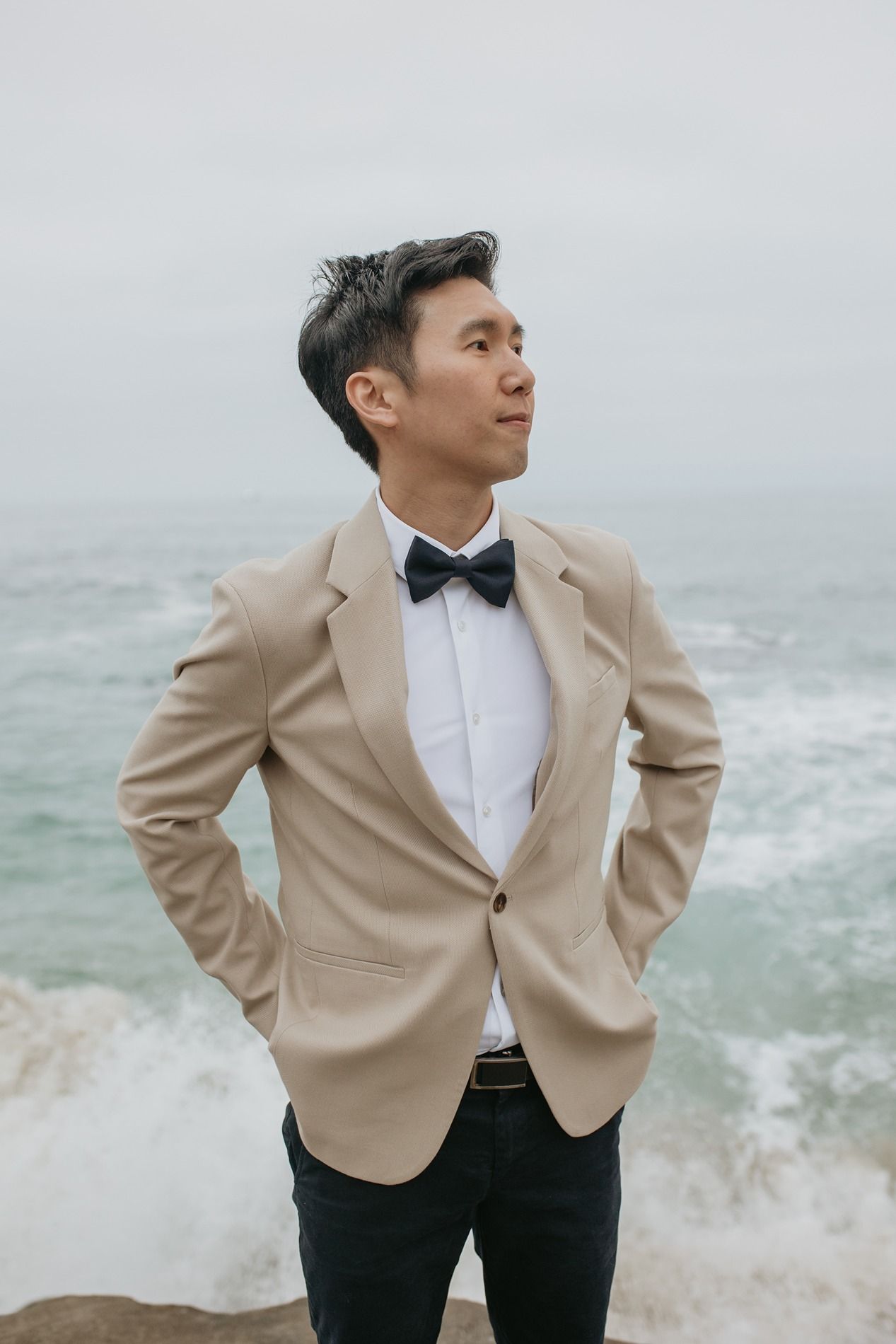 A groom in beige blazer and black pants poses against misty ocean backdrop at Windansea Beach in La Jolla.