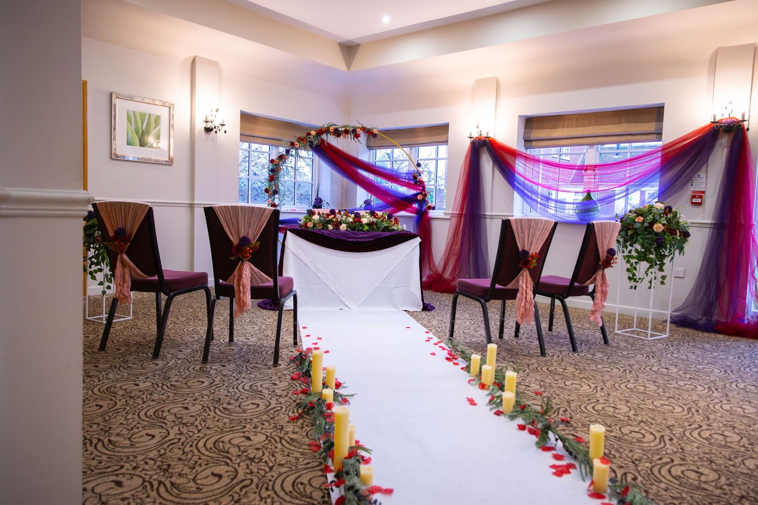 Panoramic view of an elegant event space with purple draping, white aisle runner and floral decorations along the chairs.