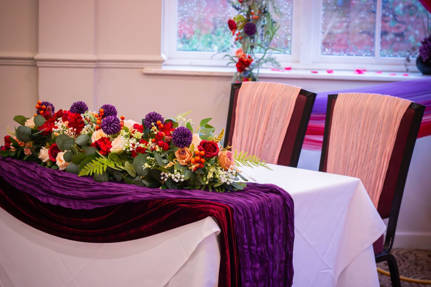 Pink dining chairs and floral centerpieces decorate an elegant banquet hall with purple accent lighting.