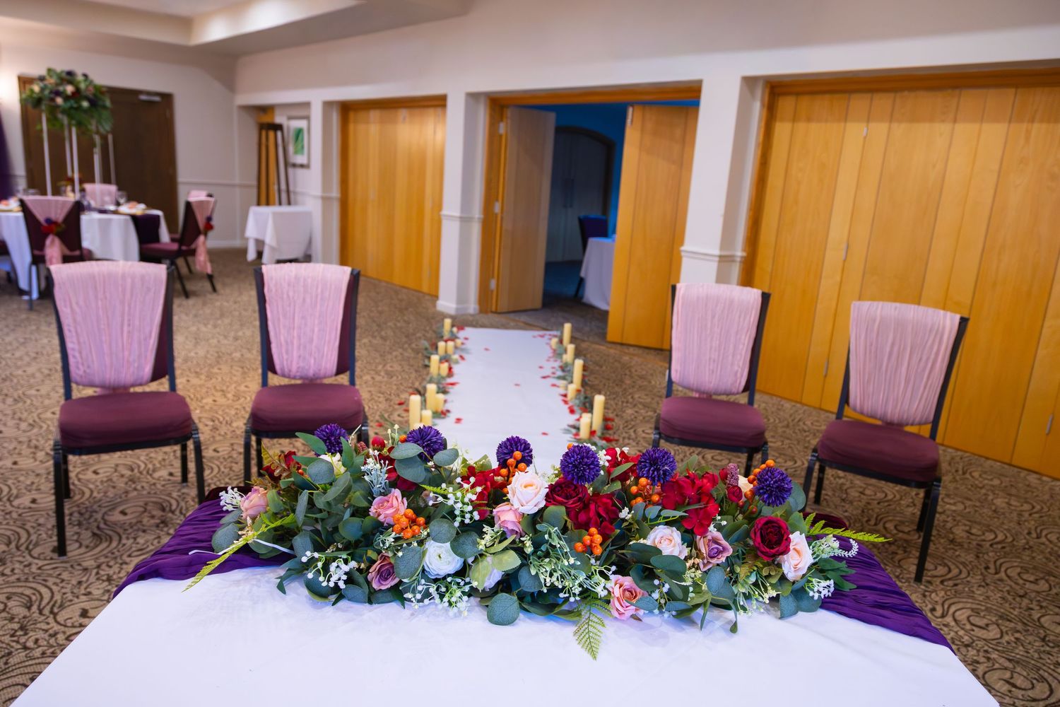 Event hall setup with purple chairs and colorful floral arrangements lining the aisle for a ceremony.