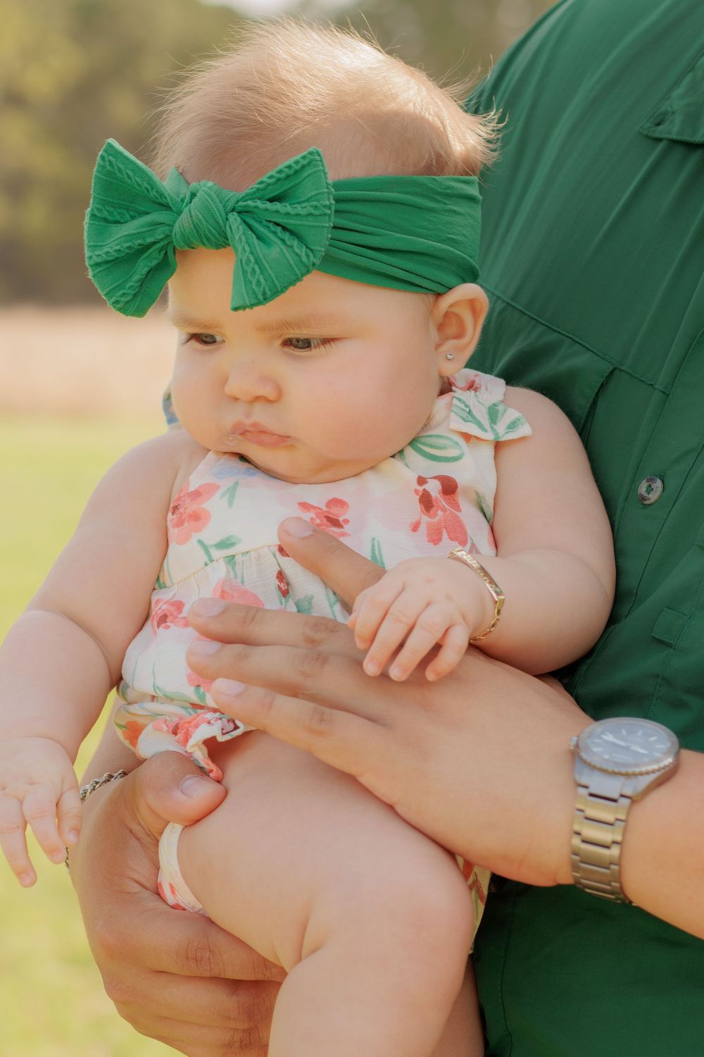 A baby wearing a floral romper and green bow headband is held outdoors.
