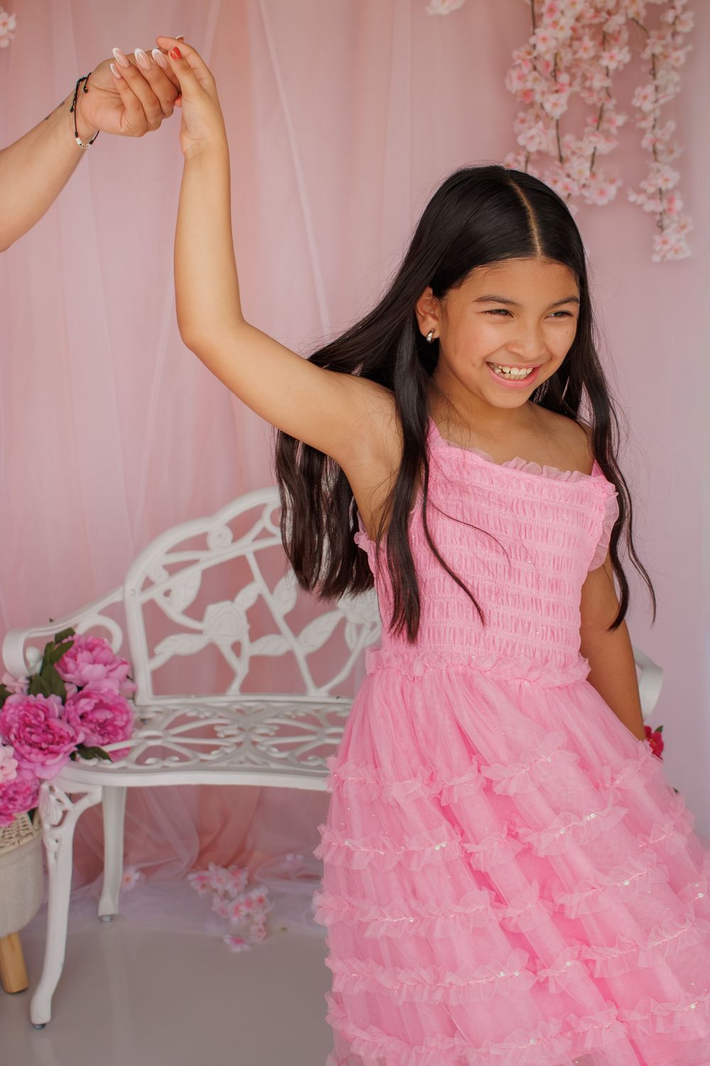 A smiling young girl in a pink ruffled dress dances in a pink floral studio with a white decorative bench.