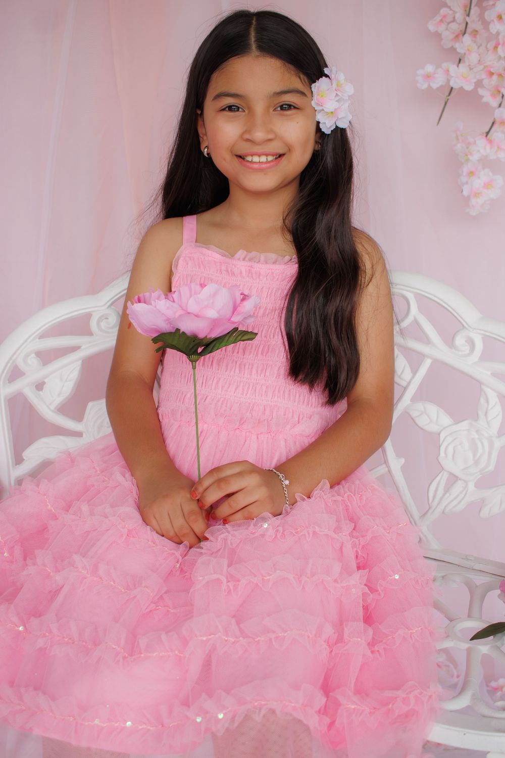 Young girl in pink ruffle dress holding a pink rose, wearing flower hair clip, smiling in a pink floral studio setting.