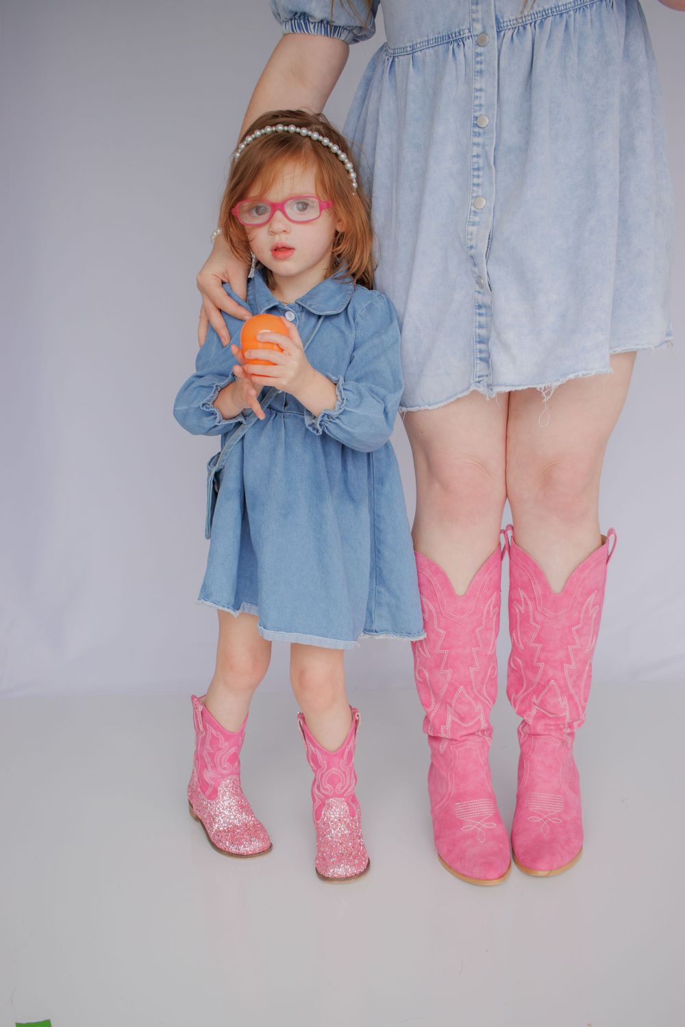 A young girl wearing a denim dress and pink cowboy boots stands in front of a white background.