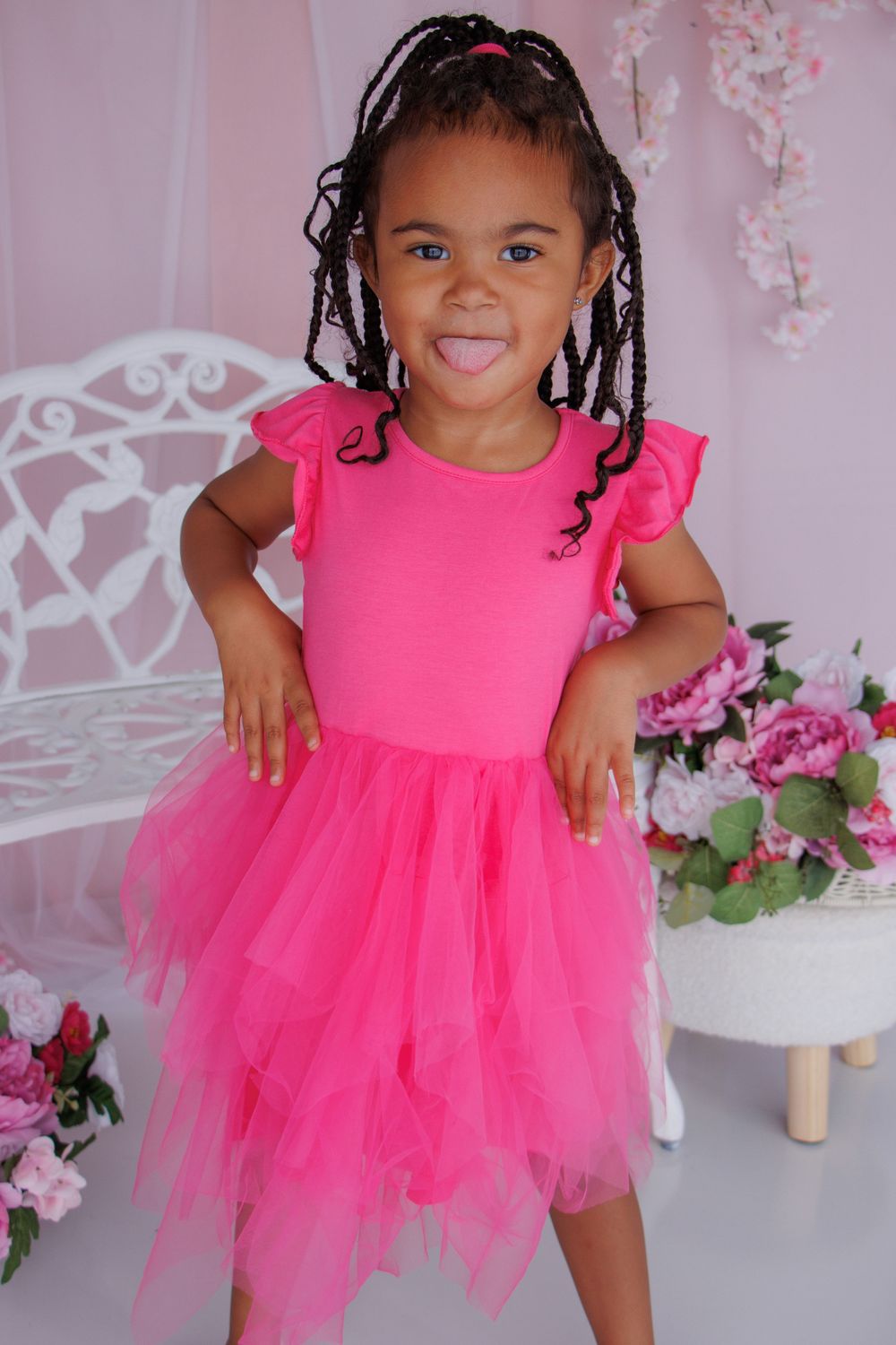 Young girl in bright pink tutu dress with braids, posing playfully in a pink floral studio setting.