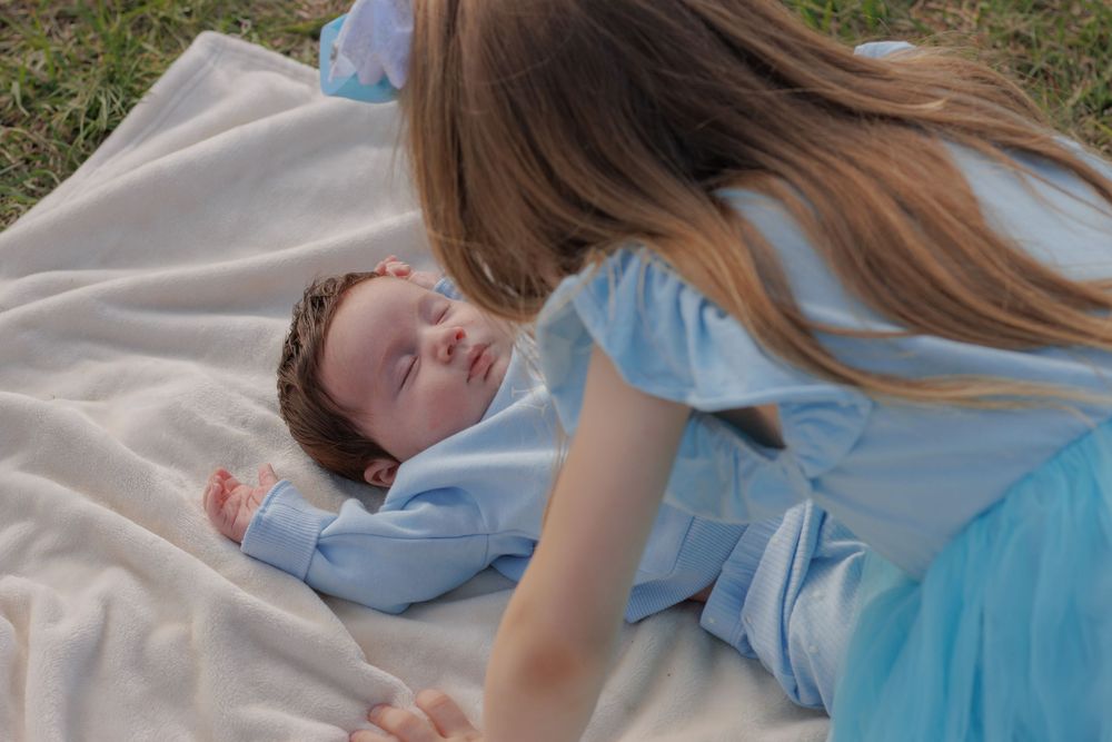 A woman lying on a blanket with a sleeping newborn baby in a grassy outdoor area.