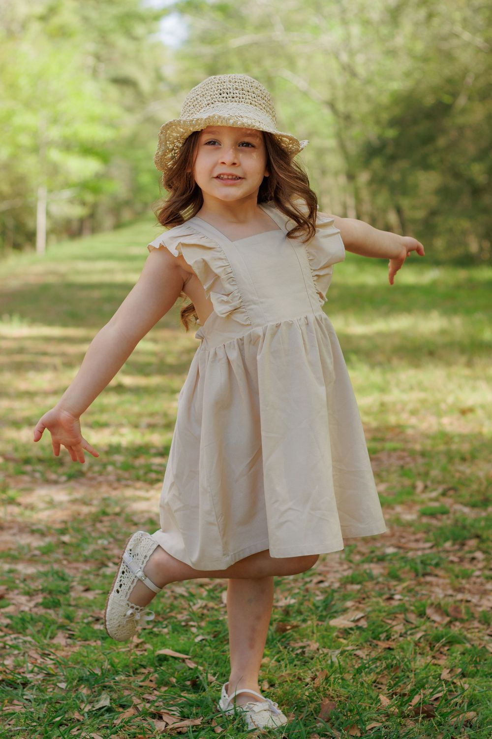 A young girl wearing a beige dress and a straw hat stands in a grassy field surrounded by trees.