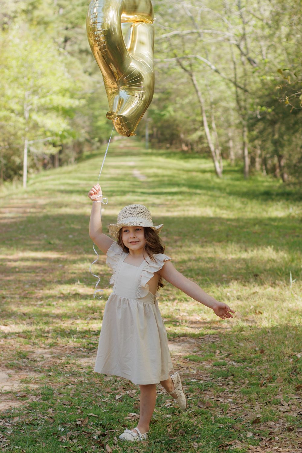 A young girl in a white dress and hat stands in a forest path, holding a gold number 5 balloon.