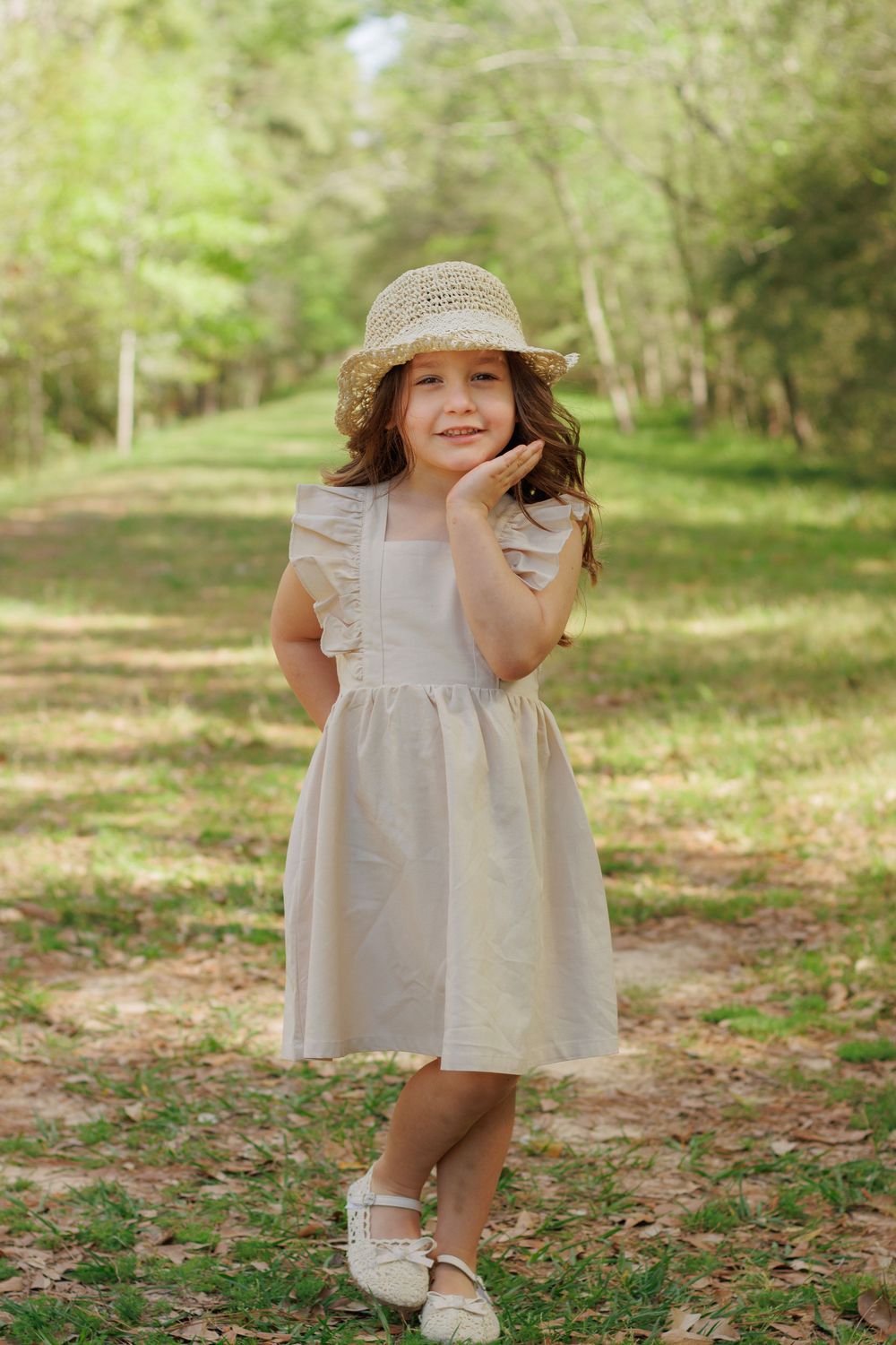 A young girl wearing a straw hat and a light-colored dress stands in a wooded area with green trees and foliage.